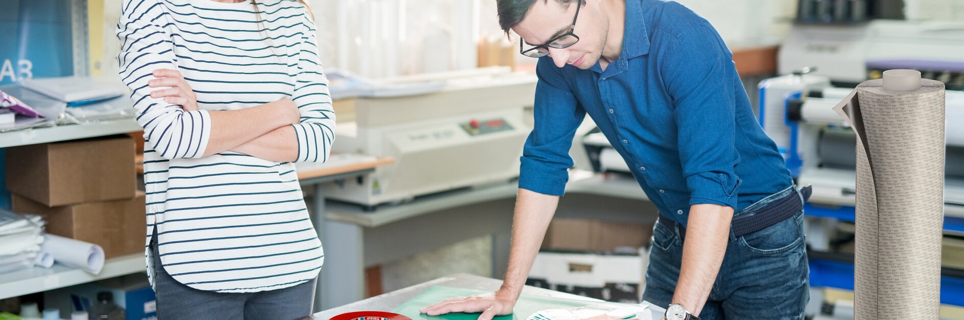 Two people in a print shop examine a large poster on a table. The man is wearing glasses and a blue shirt, while the woman is in a striped shirt with her arms crossed. The table is cluttered with rolled-up paper, tesa tape, and a laptop. (This text has been generated by AI)