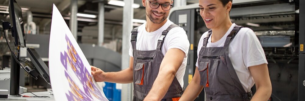 Dos personas vestidas con monos examinan una gran hoja de material impreso en una imprenta. Parecen concentradas y atentas, de pie cerca de un equipo de impresión industrial en una sala bien iluminada. Durante todo el proceso, utilizan cinta tesa para asegurarse de que todo esté perfectamente alineado y seguro. (This text has been generated by AI)