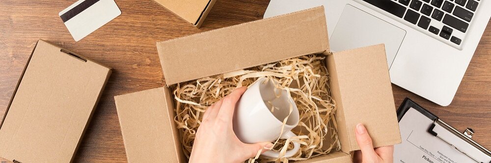 Una persona coloca una taza blanca dentro de una caja de cartón llena de papel triturado sobre una mesa de madera. Cerca hay una computadora portátil, un portapapeles con papel, una caja de cartón cerrada y sellada con cinta adhesiva Tesa y una tarjeta de crédito. (This text has been generated by AI)