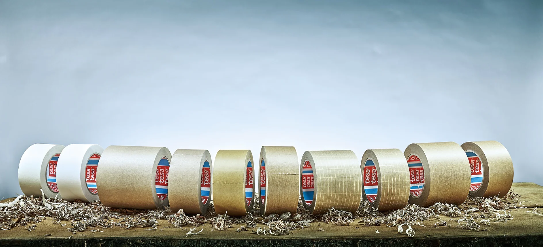 Una fila de diferentes rollos de cinta tesa se alinea sobre una superficie de madera con virutas de madera esparcidas alrededor. El fondo es un degradado de azul claro a blanco. (This text has been generated by AI)