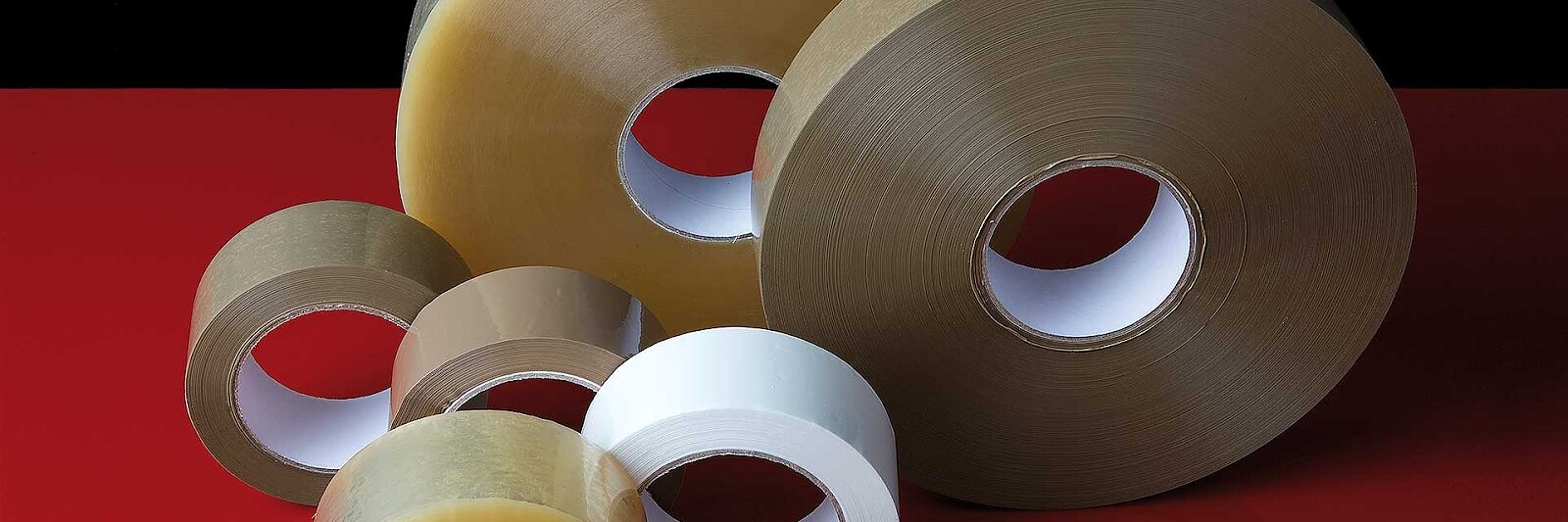 Varios rollos de cinta tesa de distintos tamaños y colores, incluidos transparente, marrón y blanco, están dispuestos sobre una superficie roja y negra. Cada rollo tiene un núcleo de cartón visible. (This text has been generated by AI)