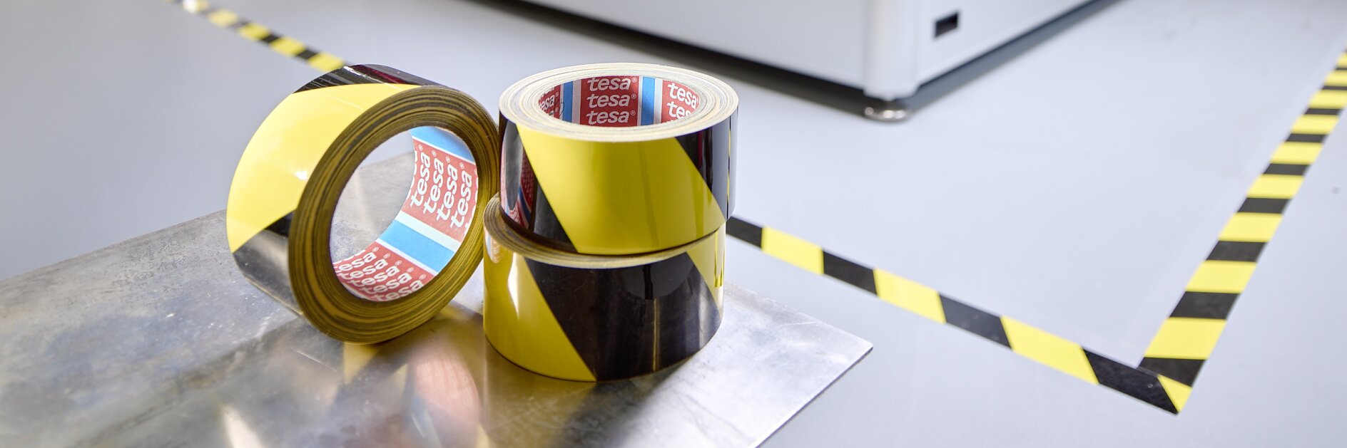 A large industrial machine inside a factory setting, with two rolls of yellow and black striped tesa tape in the foreground on the floor. The area around the machine is marked with similar caution tesa tape.