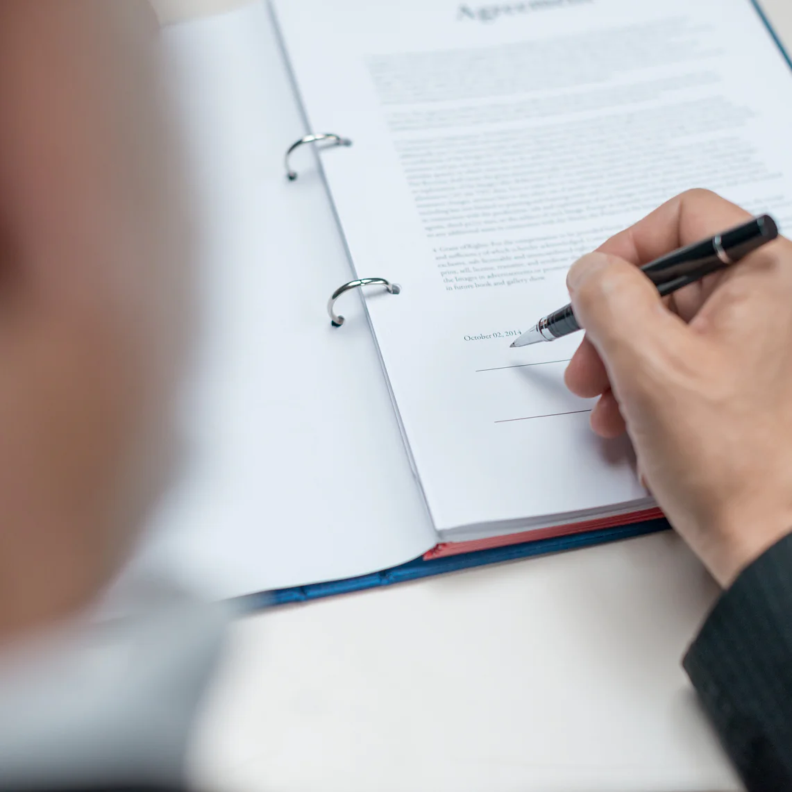 Una persona con traje sostiene un bolígrafo y se dispone a firmar un documento titulado Acuerdo. El documento está colocado en una carpeta, asegurada con cinta adhesiva, abierta en una página con texto y una línea para la firma en la parte inferior. El foco está puesto en la mano y el documento. (This text has been generated by AI)