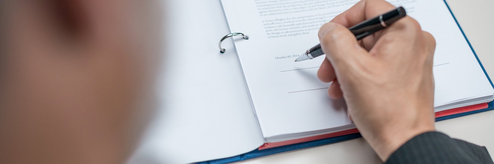Una persona con traje sostiene un bolígrafo y se dispone a firmar un documento titulado Acuerdo. El documento está colocado en una carpeta, asegurada con cinta adhesiva, abierta en una página con texto y una línea para la firma en la parte inferior. El foco está puesto en la mano y el documento. (This text has been generated by AI)