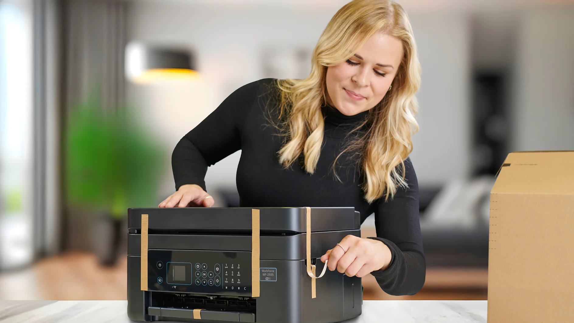 Appliance paper-strapping tape used as transport security on copierprinter Una mujer de pelo largo y rubio que lleva un jersey negro instala una impresora negra sobre una mesa de color claro. Está quitando la cinta adhesiva Tesa de la impresora. A la derecha se ve una caja de cartón y al fondo se ve una sala de estar borrosa. (This text has been generated by AI)