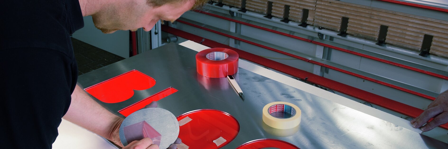 Una persona está recortando cuidadosamente letras de vinilo rojo sobre una lámina metálica con una herramienta de precisión. Sobre la mesa hay dos rollos de cinta tesa roja y un rollo de cinta tesa transparente. Junto a los rollos hay una herramienta de corte y en el fondo se ven líneas rojas.