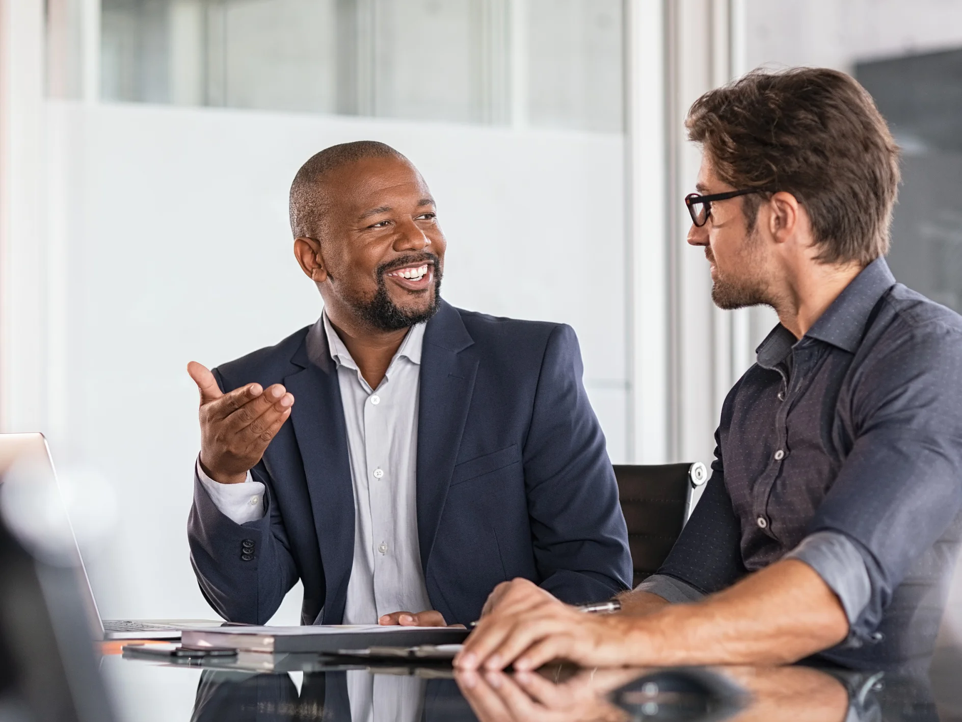 Two happy business colleagues at meeting in modern office interior. Successful african boss in a conversation with young employee in boardroom. Marketing team of two businessmen discussing strategy in meeting room.