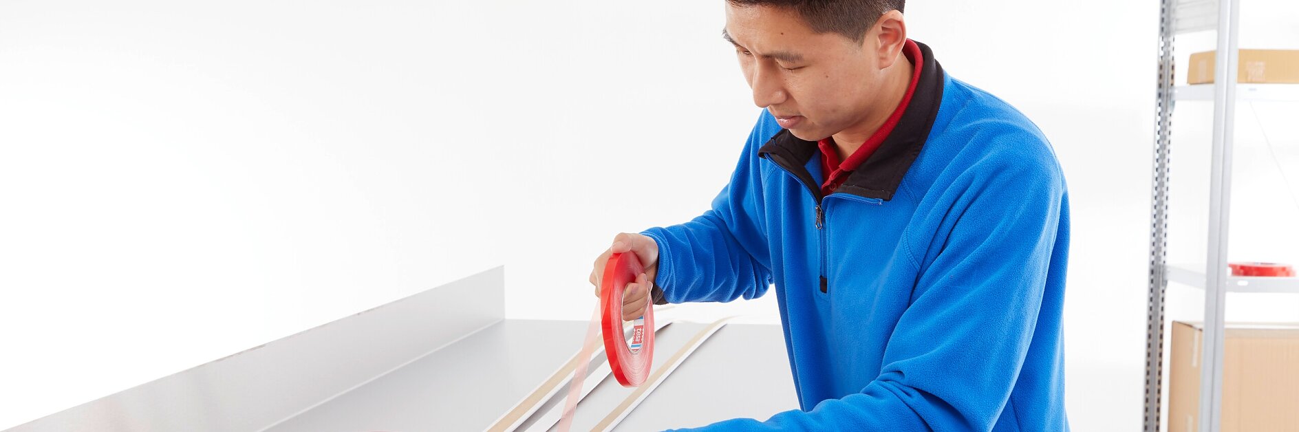 Una persona con chaqueta azul aplica cinta adhesiva roja de doble cara Tesa a listones de madera sobre una mesa de trabajo metálica. Al fondo se ven dos rollos más de cinta Tesa y un estante con cajas. (This text has been generated by AI)