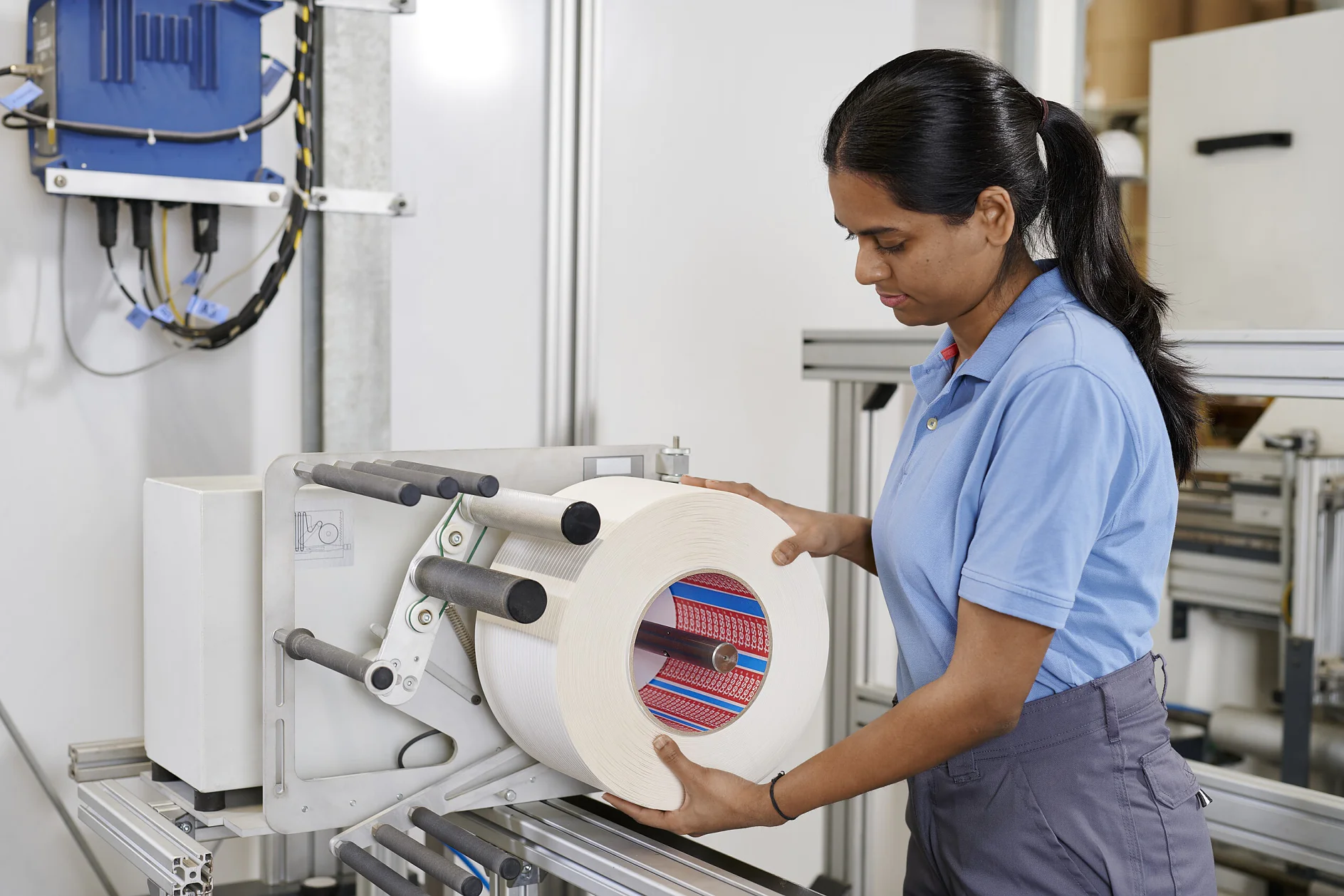 woman in blue tesa shirt operating a machine with a large roll of material in an industrial indoor setting (This text has been generated by AI)