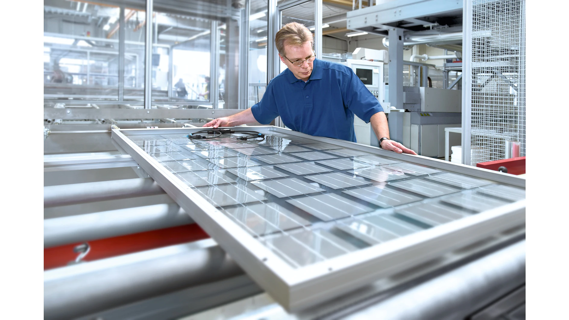Un hombre con gafas de seguridad y camisa azul comprueba un gran panel solar cerca de cinta tesa en una línea de producción de una moderna fábrica. (This text has been generated by AI)