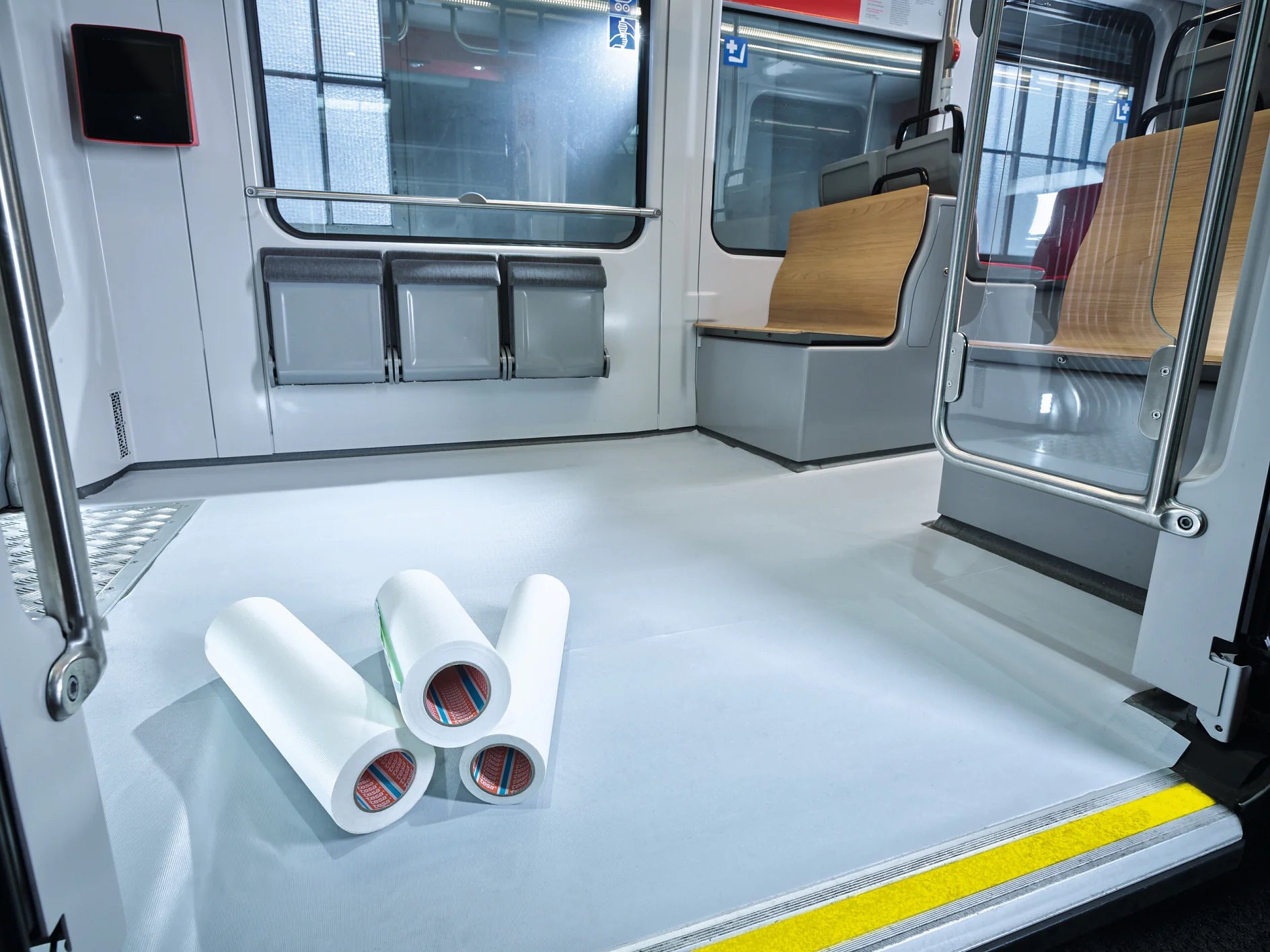 tesa-4416-cinta-antideslizante-imagen-005 Tres grandes rollos de cinta tesa blanca están en el suelo de un vagón de tren vacío y moderno, con asientos de madera y grises y una línea de seguridad amarilla. (This text has been generated by AI)