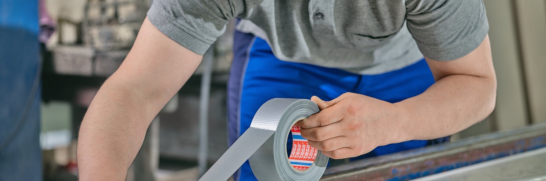tesa worker in gray shirt and blue pants applying silver duct tape to a large flat surface in an indoor workshop setting