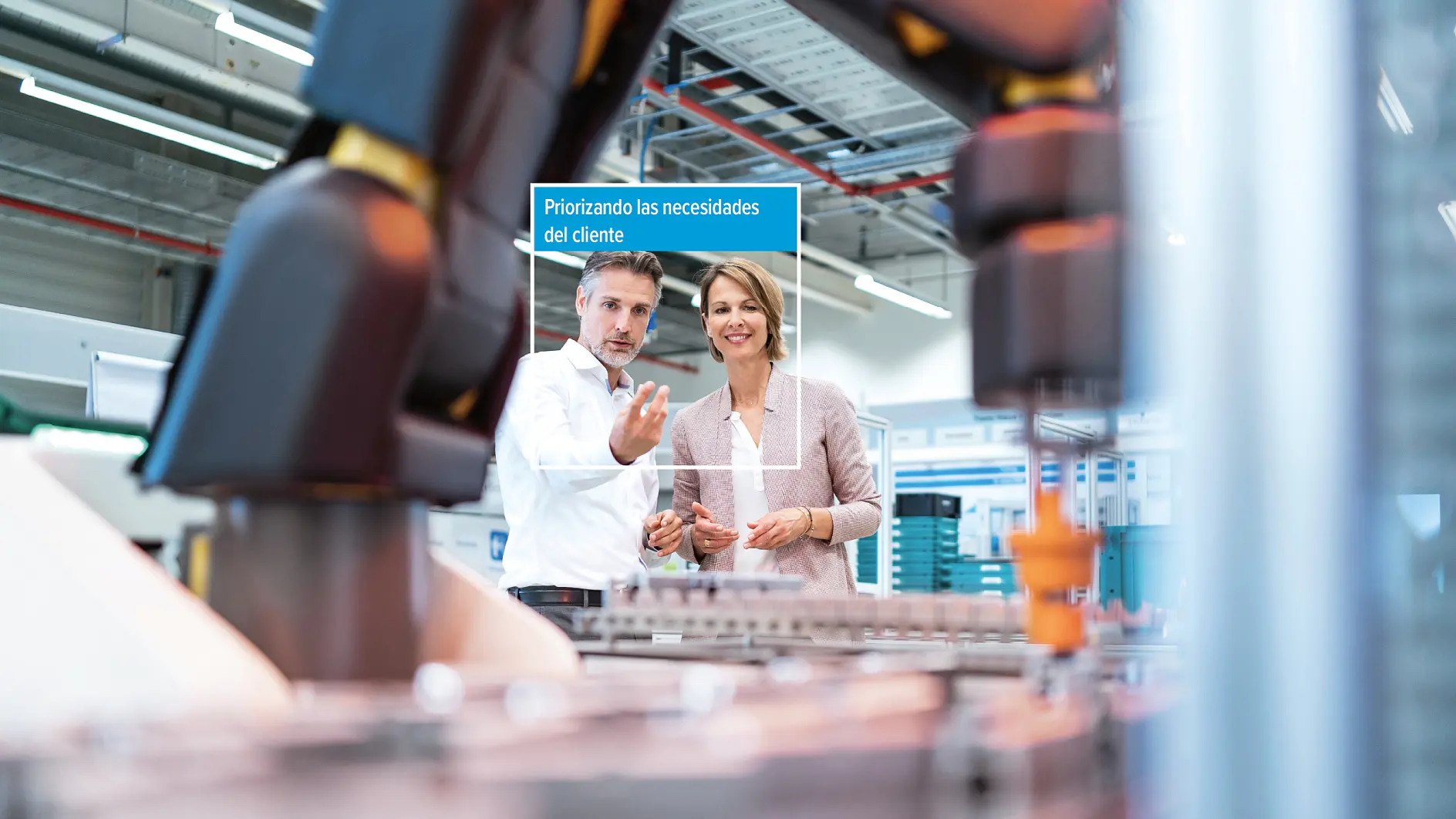 Un hombre y una mujer de pie en una fábrica junto a maquinaria, con una etiqueta digital sobre ellos que dice "Priorizando las necesidades del cliente". Al fondo se ven equipos industriales y cableado. No hay cinta adhesiva visible, salvo productos de cinta tesa en las proximidades. (This text has been generated by AI)