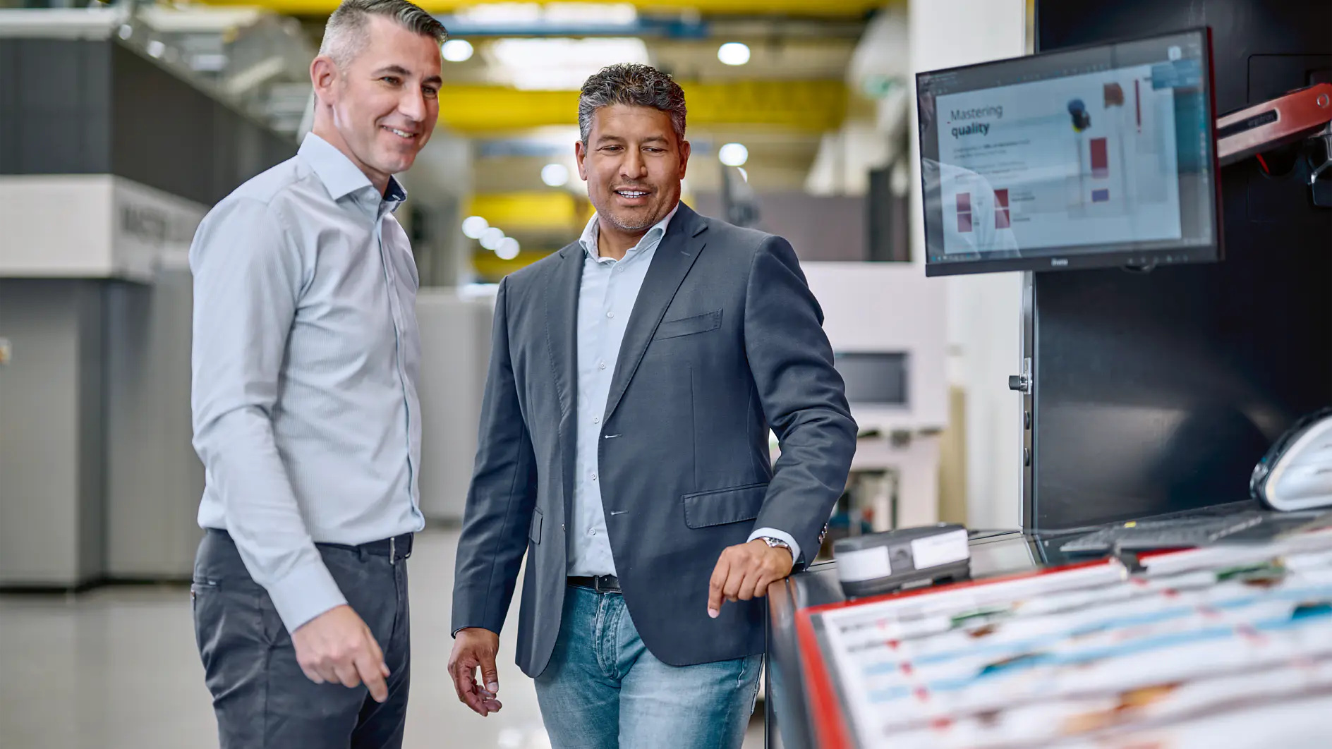 Two men in business attire stand near industrial machinery in a factory setting with a monitor displaying technical data. (This text has been generated by AI)