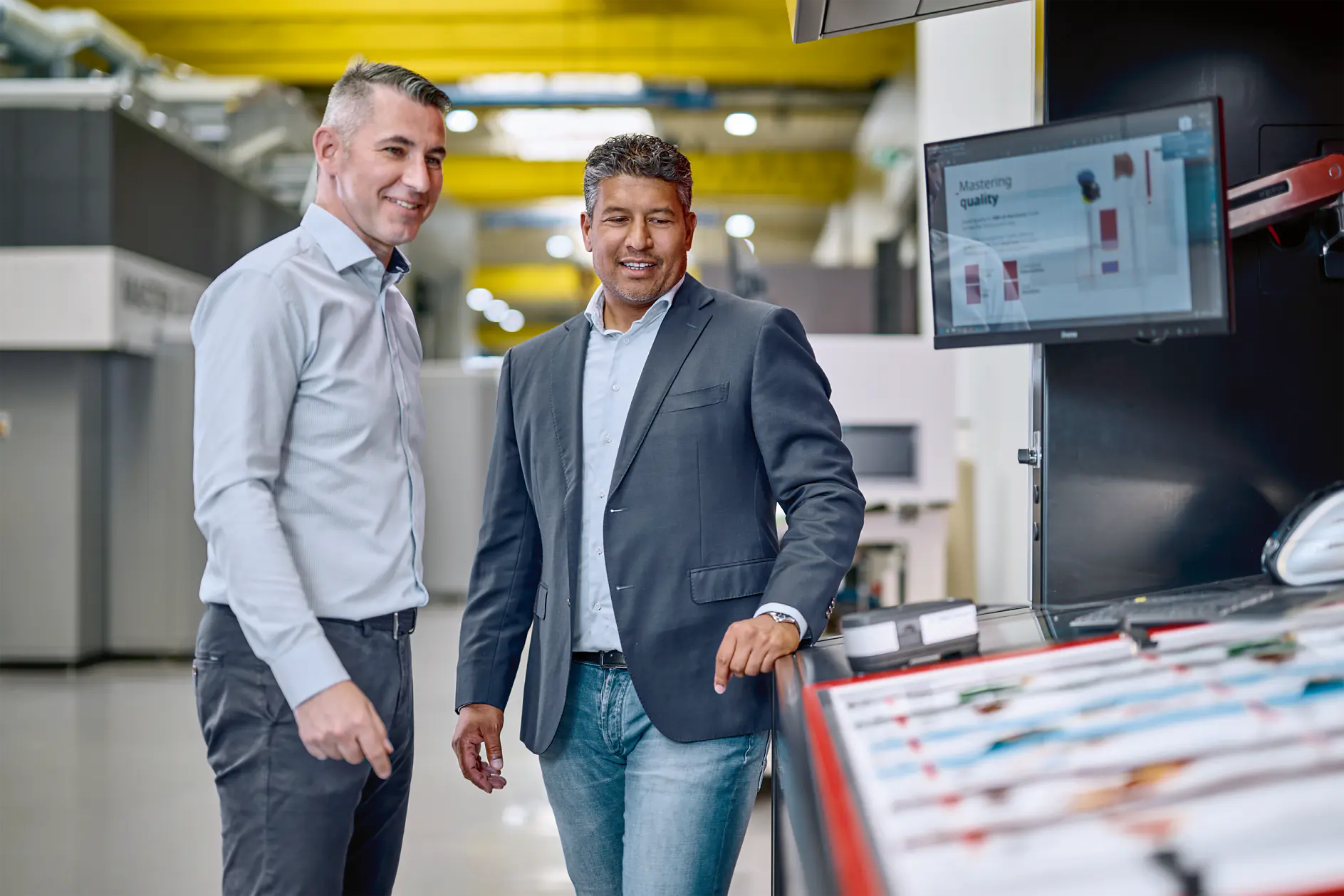 Two men in business attire stand near industrial machinery in a factory setting with a monitor displaying technical data. (This text has been generated by AI)