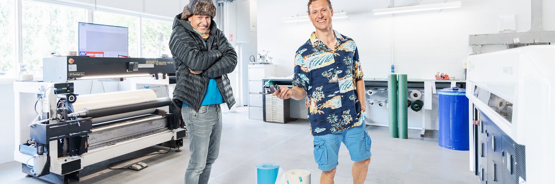 Dos personas están en una sala industrial iluminada. Una lleva chaqueta y jeans, y la otra lleva una camisa floreada, shorts y sandalias. Están de pie cerca de un equipo de impresión grande y rollos de papel. En el fondo hay diversas herramientas, maquinaria y rollos de cinta tesa.