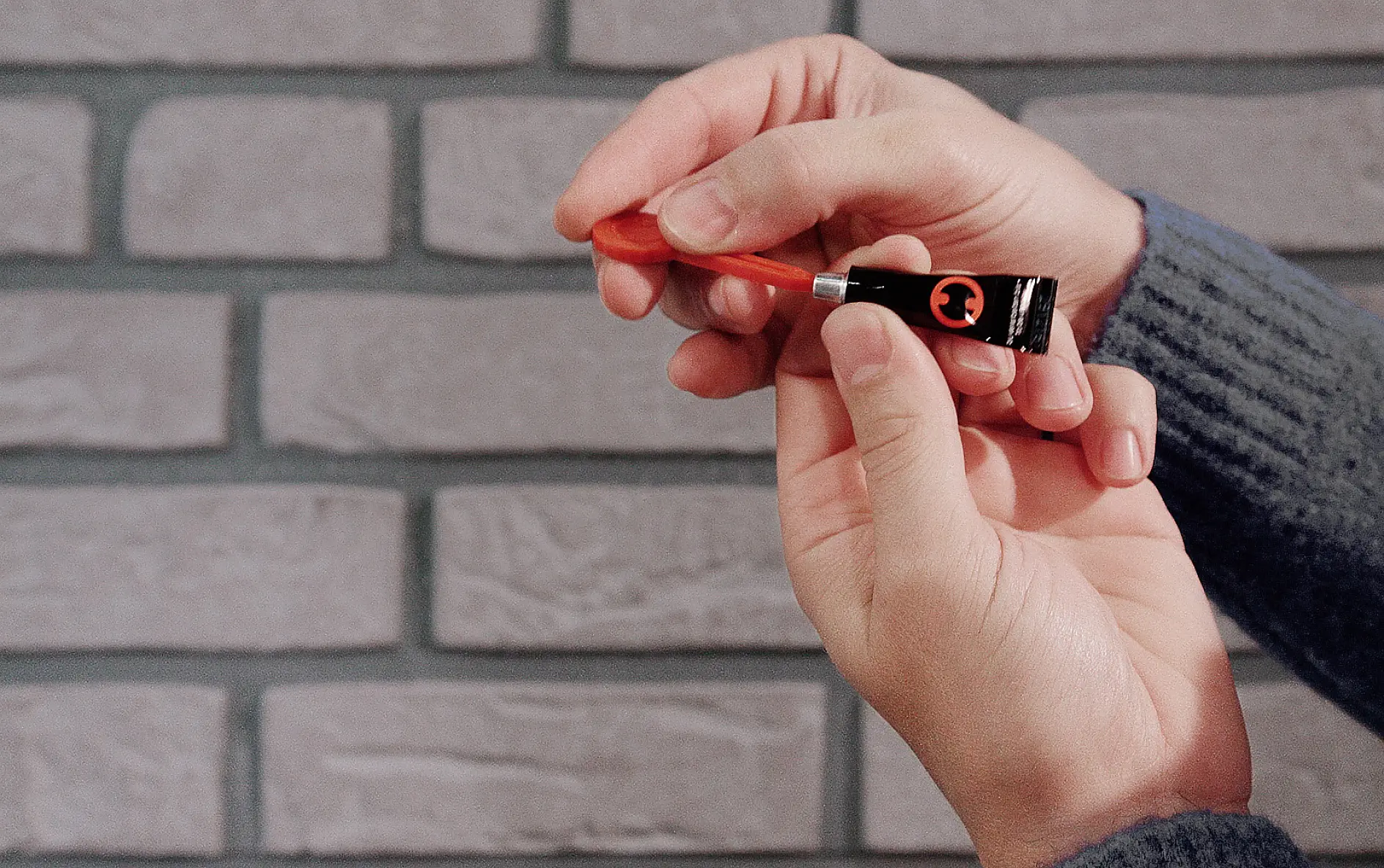 A person holding a small black tube of paint in one hand and a thin paintbrush in the other, applying paint to the brushs tip. The background features a gray brick wall with strips of tesa tape neatly positioned for guidance. (This text has been generated by AI)