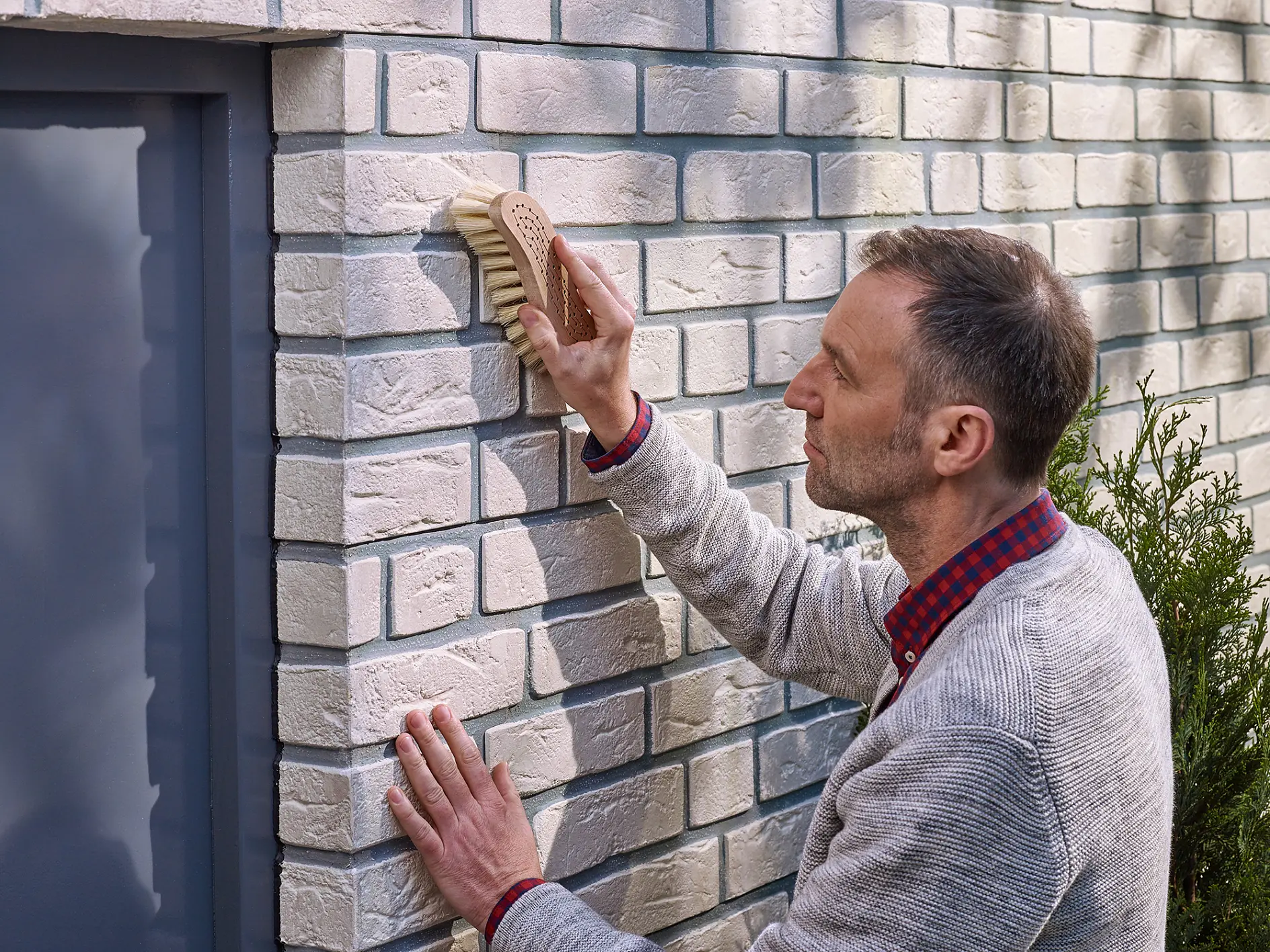 A person with short hair uses a brush to clean a white brick wall. They are wearing a gray sweater over a red plaid shirt and are positioned next to a gray door. A bush is visible in the foreground. (This text has been generated by AI)