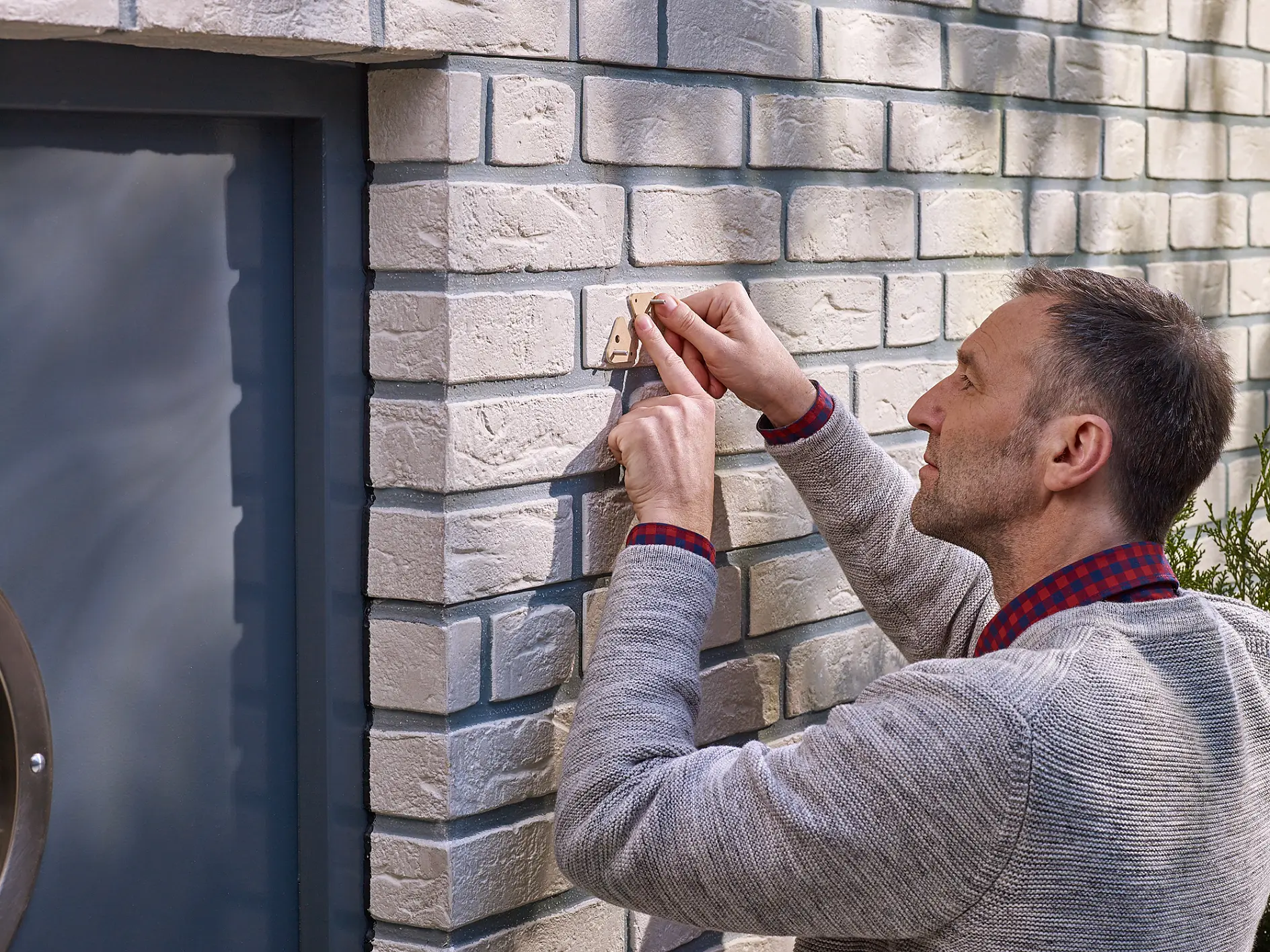 A man in a gray sweater and checkered shirt installs a smart doorbell on a white brick wall near a blue door. He appears focused on attaching the device securely to the wall using tesa tape. (This text has been generated by AI)