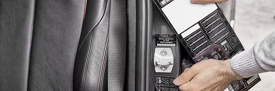 Una persona coloca una etiqueta con un código de barras y texto en el marco interior de la puerta de un automóvil, cerca de una bisagra, utilizando cinta adhesiva Tesa. El interior del automóvil es negro con costuras rojas. Se ven varias etiquetas en el marco de la puerta. (This text has been generated by AI)