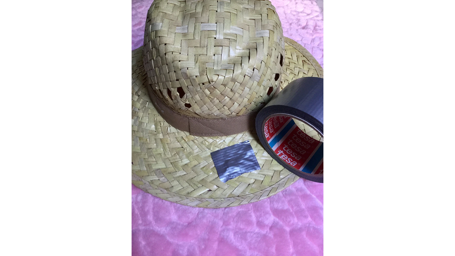 Un sombrero de paja tejido se encuentra sobre una superficie texturizada de color rosa. Junto al sombrero hay un rollo de cinta tesa gris, con un pequeño trozo de cinta tesa cortado y colocado al lado del rollo. (This text has been generated by AI)
