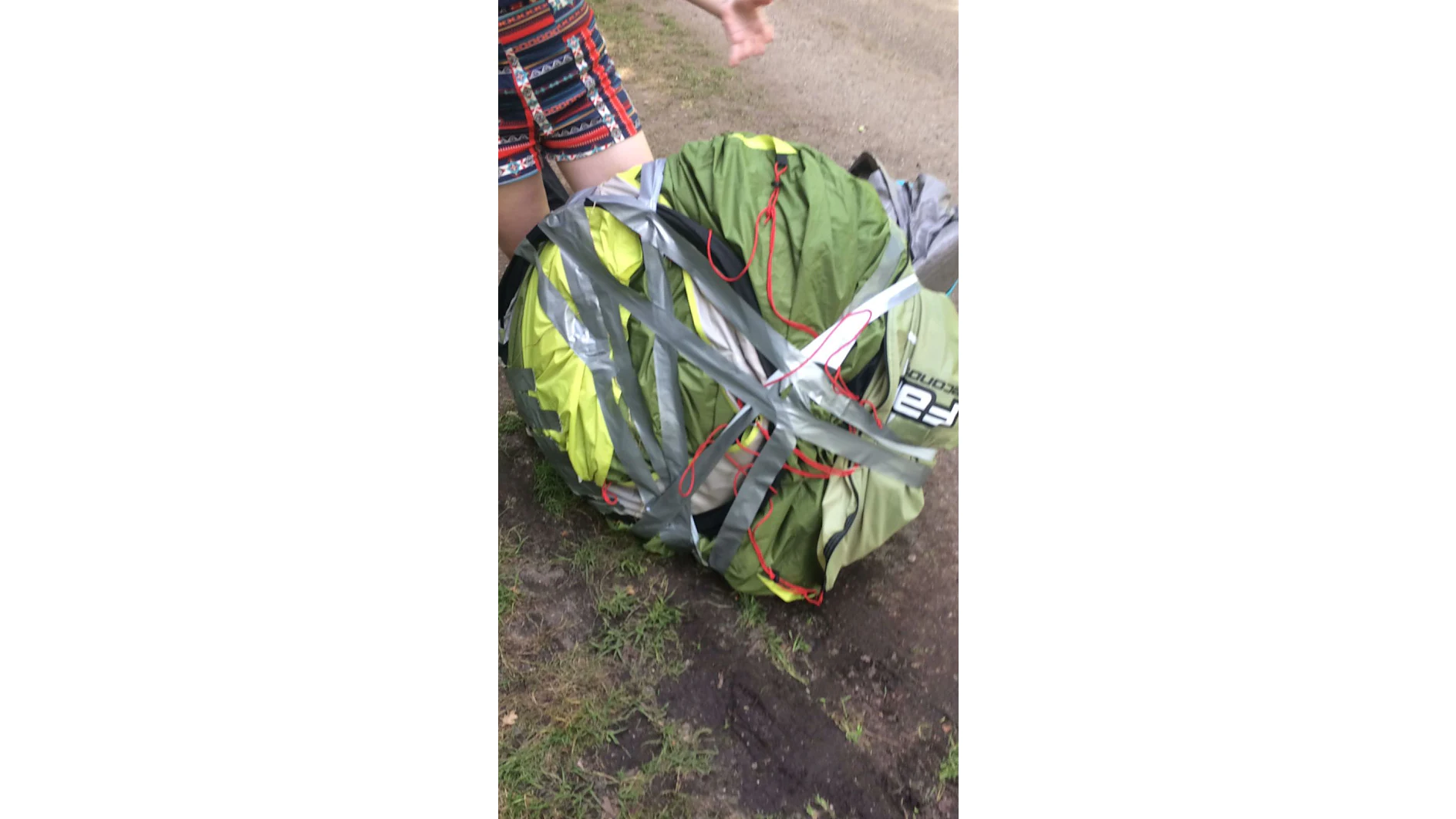 Una persona se encuentra de pie junto a una tienda de campaña verde y gris que está atada de forma desordenada y asegurada con tiras de cinta adhesiva Tesa entrecruzadas. La tienda está en el suelo rodeada de césped y un camino de tierra. La mano de la persona es parcialmente visible. (This text has been generated by AI)