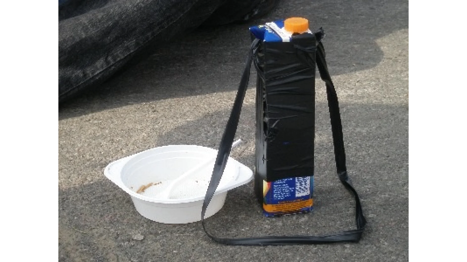 Un recipiente de plástico blanco con una cuchara reposa sobre cemento al lado de un cartón de jugo envuelto en cinta tesa negra, con la cinta tesa formando un bucle, parecido a un asa. (This text has been generated by AI)