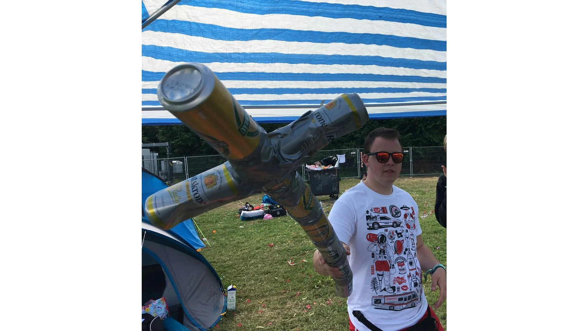 Una persona con una camiseta blanca estampada y gafas de sol sostiene un objeto en forma de cruz hecho con latas de cerveza sujetadas con cinta adhesiva bajo un toldo de rayas azules y blancas. Al fondo se ven tiendas de campaña y césped. (This text has been generated by AI)