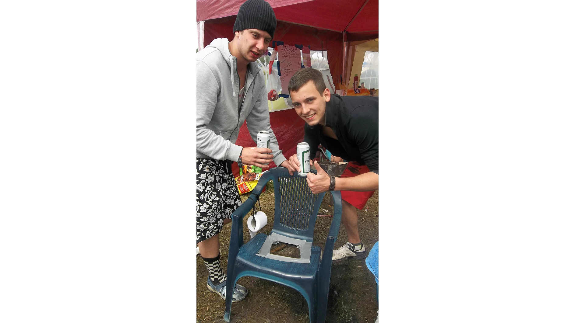 Dos hombres sonríen a la cámara mientras sostienen latas de cerveza. Están de pie junto a una silla de plástico con el asiento roto y reparaciones con cinta adhesiva Tesa. En el fondo hay un toldo rojo y un cartel de papel con una inscripción. Un hombre hace un gesto con el pulgar hacia arriba. (This text has been generated by AI)