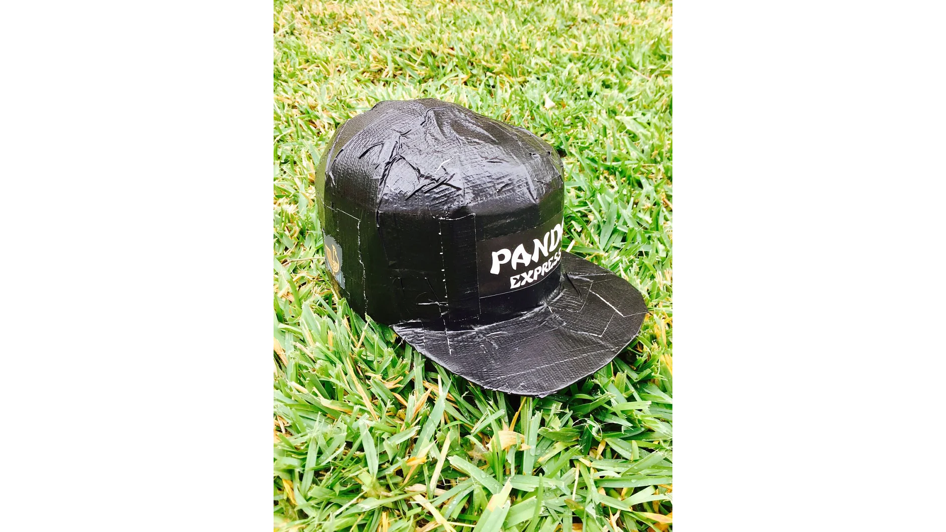 Una gorra de béisbol de Panda Express se encuentra sobre un trozo de césped verde. La gorra está completamente cubierta con cinta adhesiva tesa negra, lo que oculta su superficie pero deja visible el logotipo de la marca. (This text has been generated by AI)