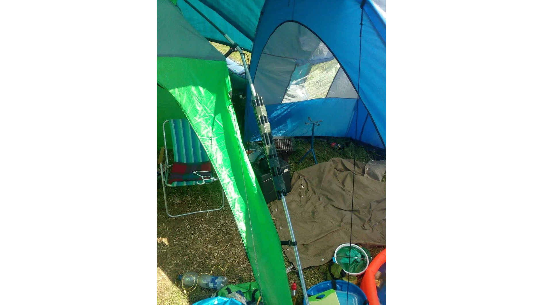 Dentro de una carpa azul y verde hay una silla plegable, una manta a rayas en el suelo, una pequeña parrilla y varios artículos de campamento dispersos, incluido un balde azul y contenedores de plástico. La carpa parece estar instalada sobre césped, sujeta de forma segura con cinta tesa para garantizar que todo esté en su lugar. (This text has been generated by AI)