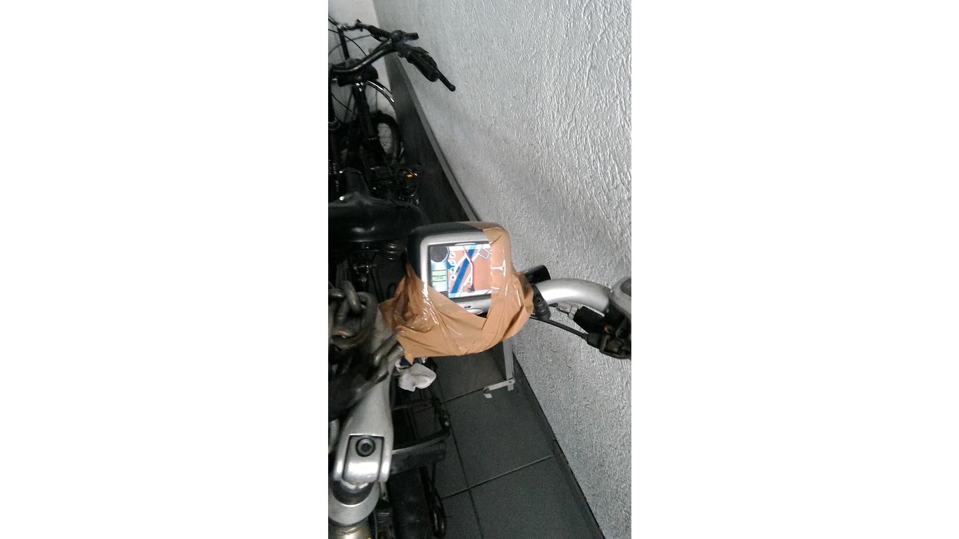 Un manillar de bicicleta con un espejo fijado con cinta adhesiva marrón Tesa. El espejo refleja una imagen de la bandera británica. Hay otras bicicletas estacionadas cerca, junto a una pared blanca con textura. (This text has been generated by AI)