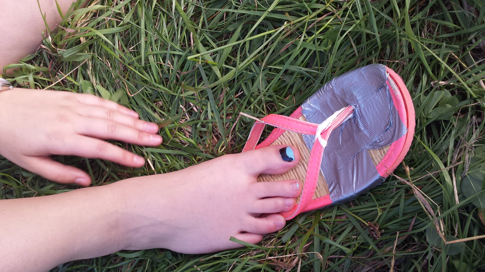 Una sandalia reparada con cinta tesa está sobre el césped. El pie de una persona con una uña pintada está medio dentro de la sandalia, con una mano extendida hacia ella. La sandalia tiene correas rosas y el pie descansa a su lado. (This text has been generated by AI)