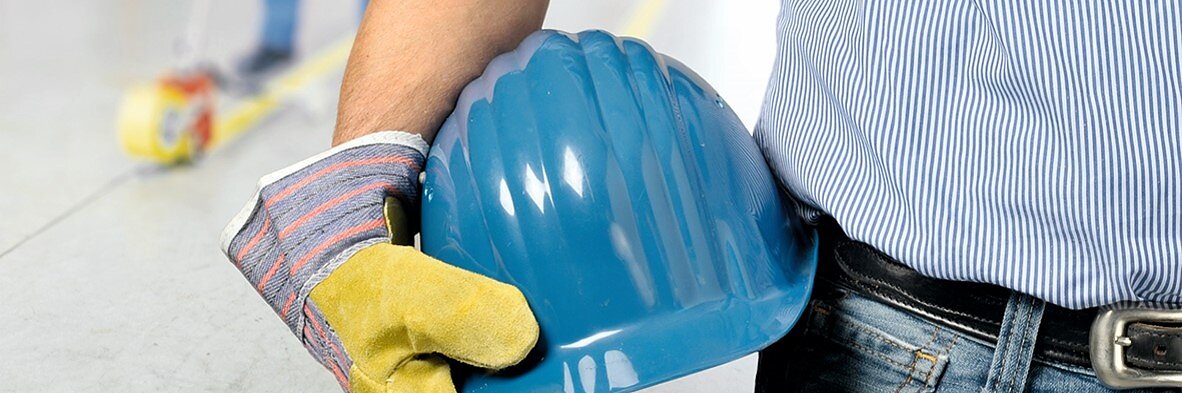 Una persona con una camisa de rayas azules y jeans sostiene un casco azul en su mano enguantada. Se encuentra de pie sobre el piso de concreto, marcado con una línea amarilla hecha con cinta tesa. Otra persona en el fondo está fregando el piso. (This text has been generated by AI)