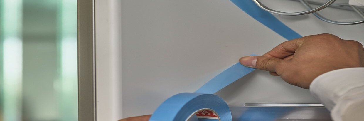 Una persona coloca una cinta adhesiva azul Tesa en el interior de un frigorífico. Sostiene el rollo de cinta Tesa en una mano y presiona la cinta sobre la superficie con la otra. El frigorífico está abierto, dejando al descubierto unos estantes metálicos y una superficie interior reflectante. (This text has been generated by AI)