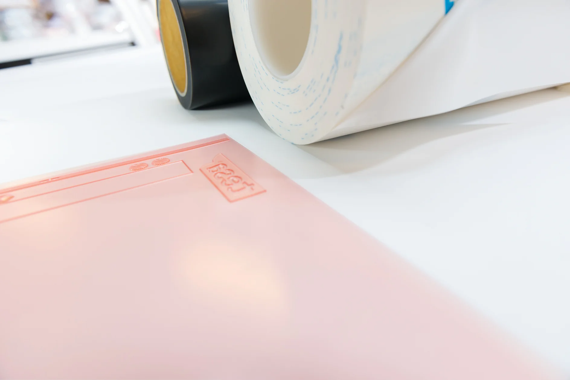 Close-up of a pink sheet with embossed text and symbols, placed on a white surface. Two rolls of tesa tape, one black and one white with blue patterns, are positioned in the background. (This text has been generated by AI)