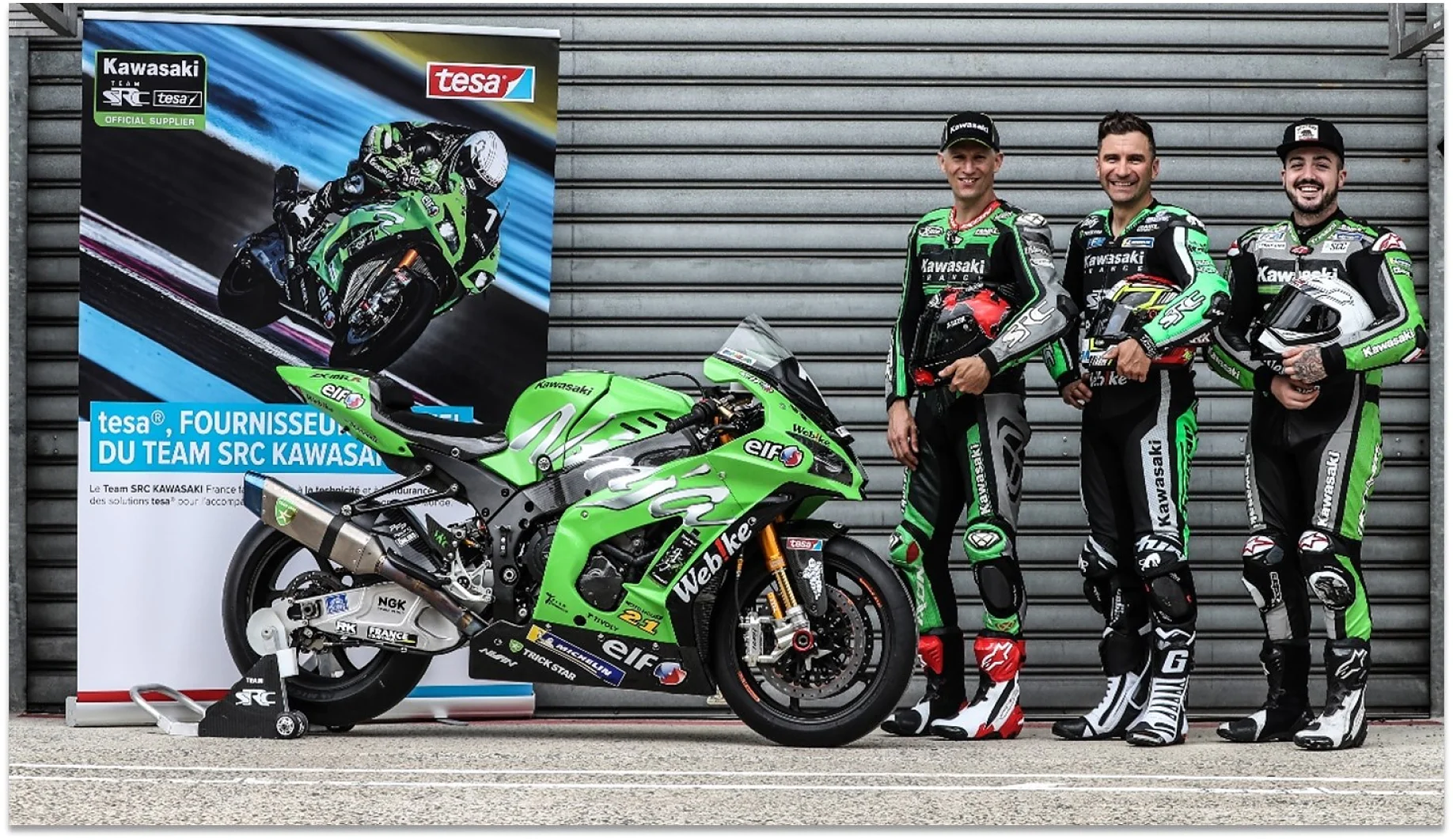 Tres motociclistas con trajes de carreras se encuentran junto a una motocicleta Kawasaki verde y negra. A la izquierda hay una pancarta con imágenes similares. Están frente a una pared de metal corrugado, con una cinta tesa que se destaca en toda la escena, lo que mejora tanto el rendimiento como el estilo. (This text has been generated by AI)