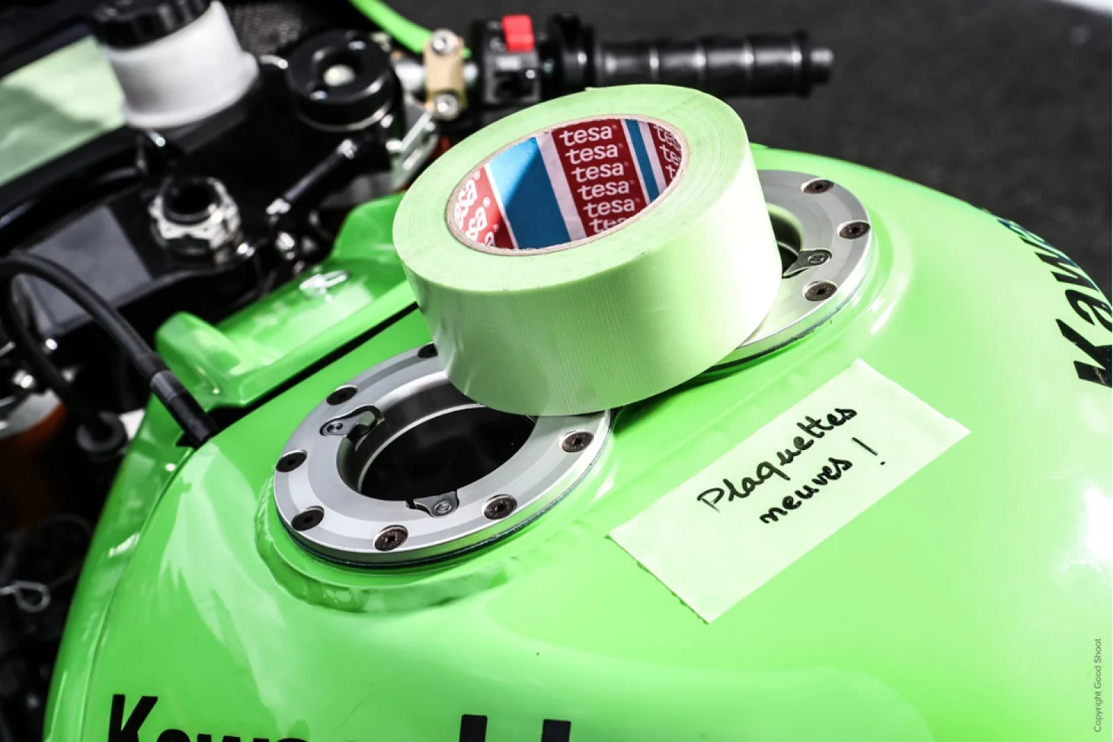 Una motocicleta verde con un tanque de combustible sobre el cual se encuentra colocado un rollo de cinta tesa verde y una nota escrita en francés que dice Plaquettes neuves!. Cerca hay un rollo de cinta tesa roja y azul. (This text has been generated by AI)