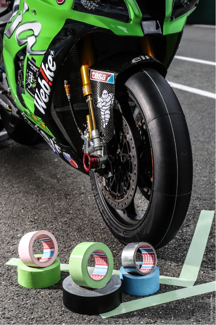 Una motocicleta con un guardabarros delantero verde está estacionada sobre el asfalto. Hay cinco rollos de cinta Tesa delante de la motocicleta, en varios colores: rosa, verde, negro, gris y azul verdoso. La cinta Tesa se aplica en líneas paralelas en el suelo. (This text has been generated by AI)