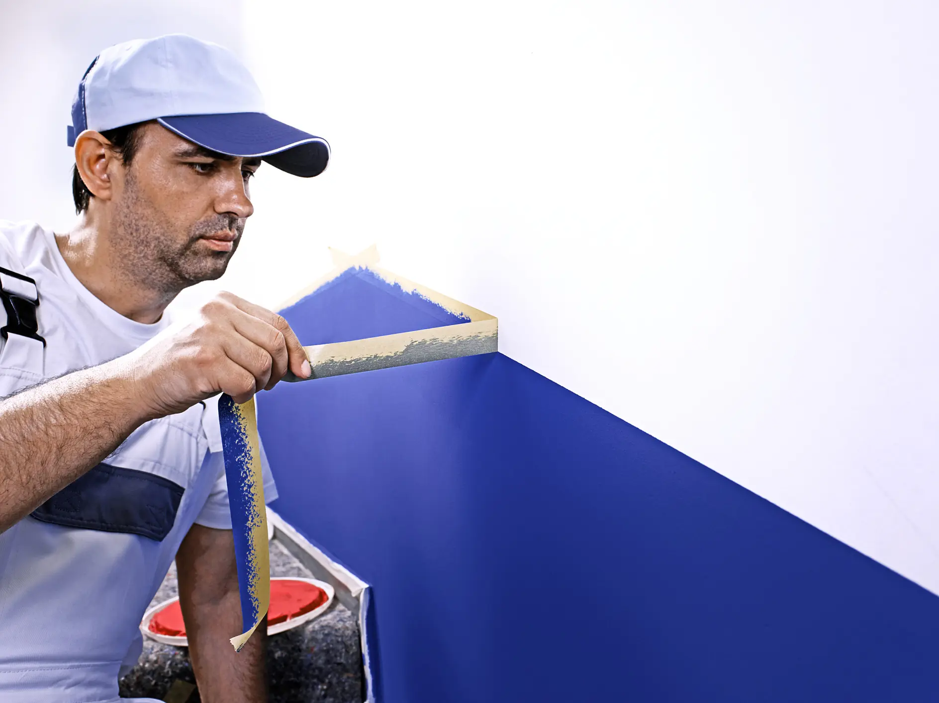 A man wearing a blue cap and a white shirt with blue overalls is painting a geometric design on a wall. He is peeling tesa tape from a blue-painted section on a white wall. A paint can is visible at his side.