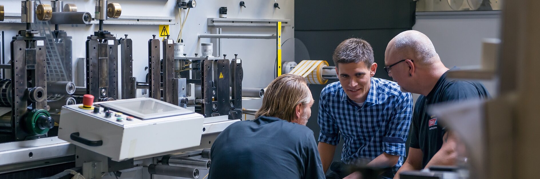 Three men are gathered around a large industrial machine in a factory setting. They appear to be engaged in discussion. The machine has various levers and components, and there are rolls of tesa tape in the background.