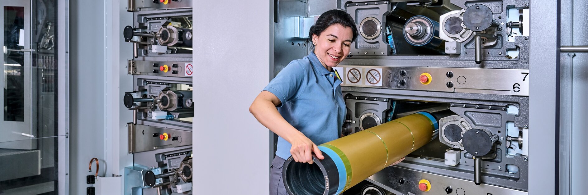 A woman in a blue polo shirt and gray pants is inserting a cylindrical part into a large industrial machine with multiple numbered sections, using tesa tape. The setting appears to be a factory or manufacturing facility. (This text has been generated by AI)