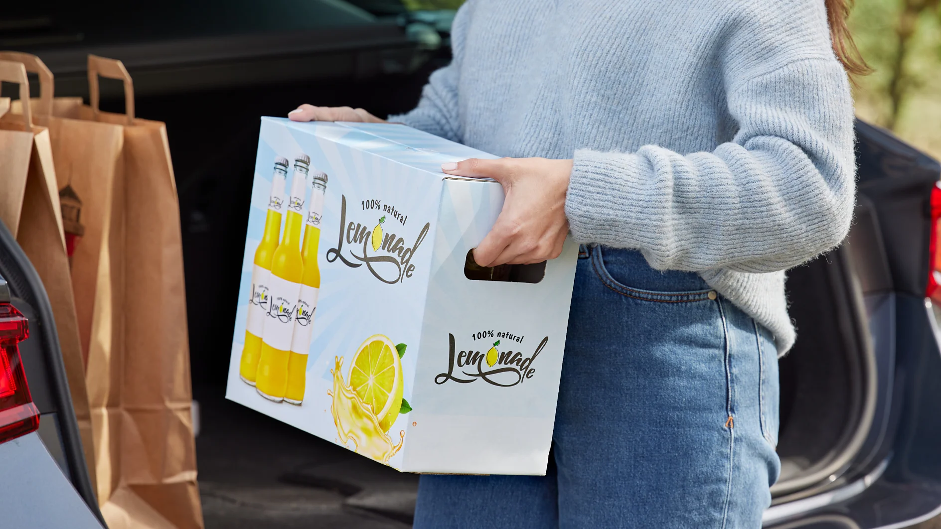 A person wearing a gray sweater and blue jeans is loading a box labeled Lemonade into the trunk of a car. The trunk also contains brown paper bags sealed with tesa tape. (This text has been generated by AI)
