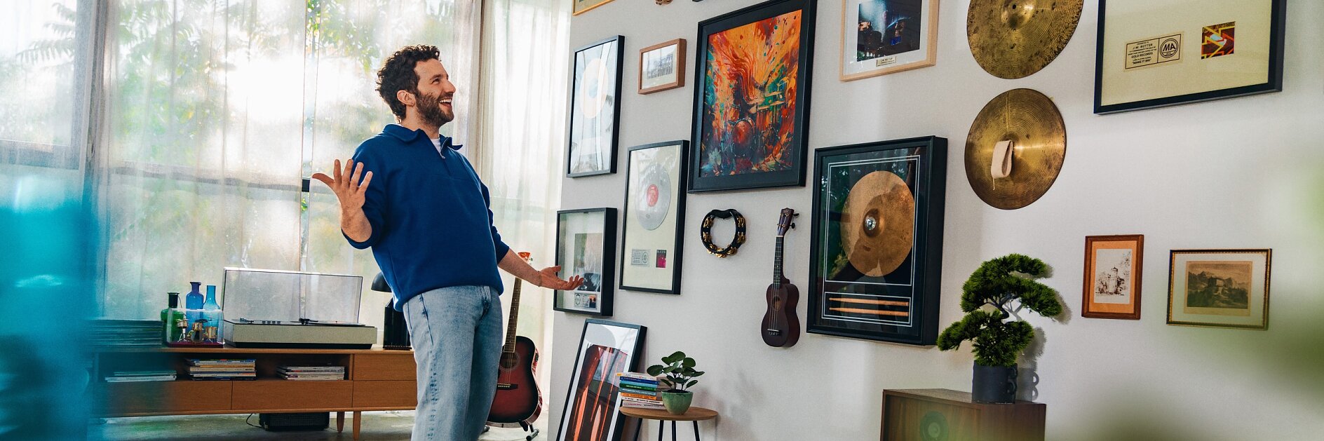 Un hombre se encuentra de pie en una habitación muy iluminada mirando una pared llena de discos, fotografías e instrumentos musicales enmarcados. Parece estar sonriendo, con los brazos ligeramente levantados. La habitación incluye un tocadiscos, altavoces y plantas frondosas. Enmarcando los discos y las fotografías en la pared hay cinta adhesiva tesa, conocida por su sujeción fiable y su acabado de calidad. (This text has been generated by AI)