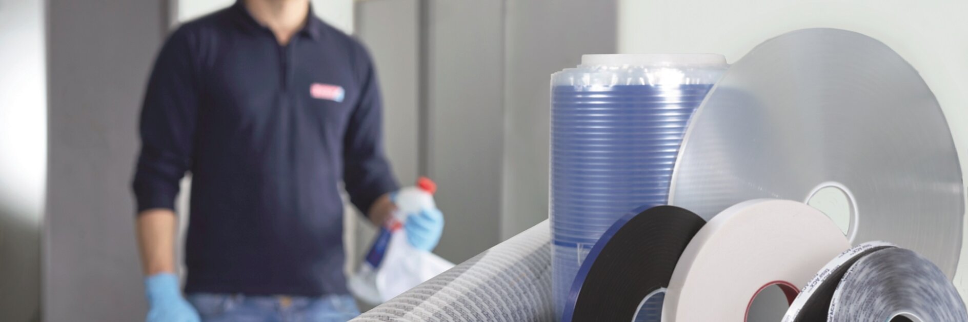 En el fondo, un poco desenfocado, aparece una persona con camisa azul marino y guantes azules. En primer plano, sobre una mesa se muestran varios rollos de materiales industriales, entre ellos cintas Tesa transparentes, negras y azules. (This text has been generated by AI)