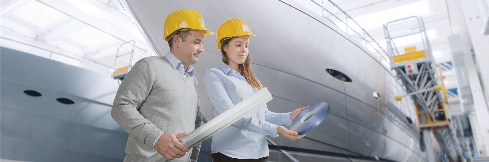 two engineers wearing yellow helmets review blueprints inside a large indoor boatyard near a white yacht under construction