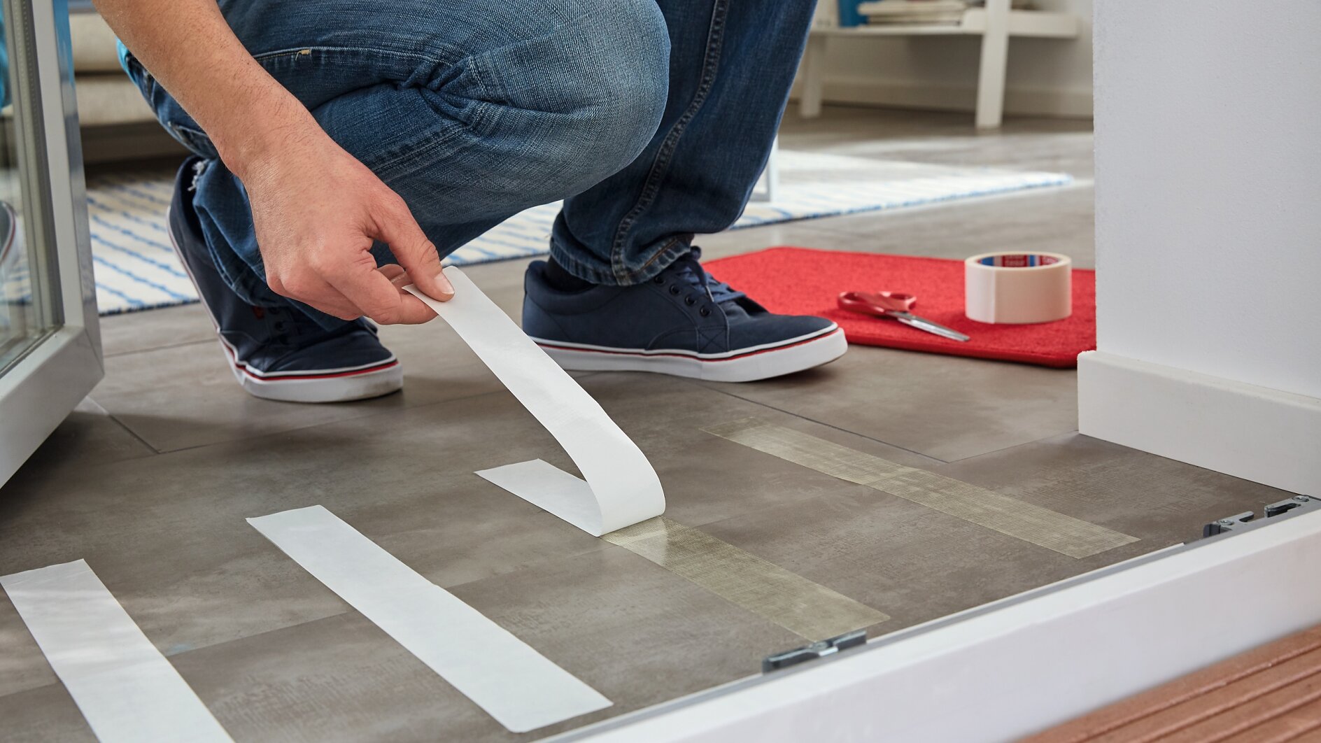 Una persona con vaqueros y zapatillas azul marino se arrodilla sobre un suelo de baldosas, aplicando tiras de cinta adhesiva de doble cara Tesa. Cerca hay un tapete rojo, tijeras y un rollo de cinta Tesa. El escenario parece ser un interior. (This text has been generated by AI)