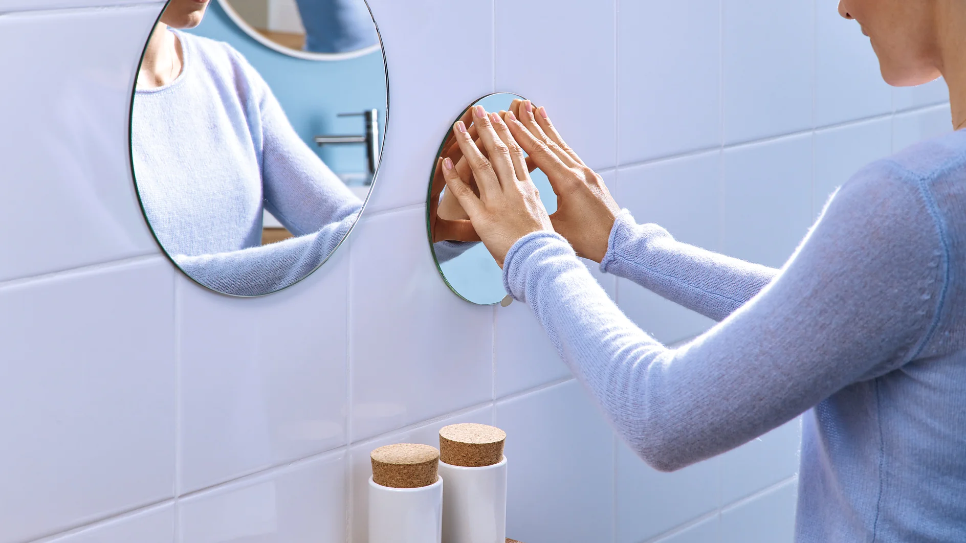 Una persona con una camisa azul claro de manga larga limpia un espejo redondo en la pared blanca de un baño con azulejos usando cinta adhesiva. Dos recipientes blancos con tapas de corcho y una toalla blanca doblada están sobre la encimera de madera de abajo. (This text has been generated by AI)