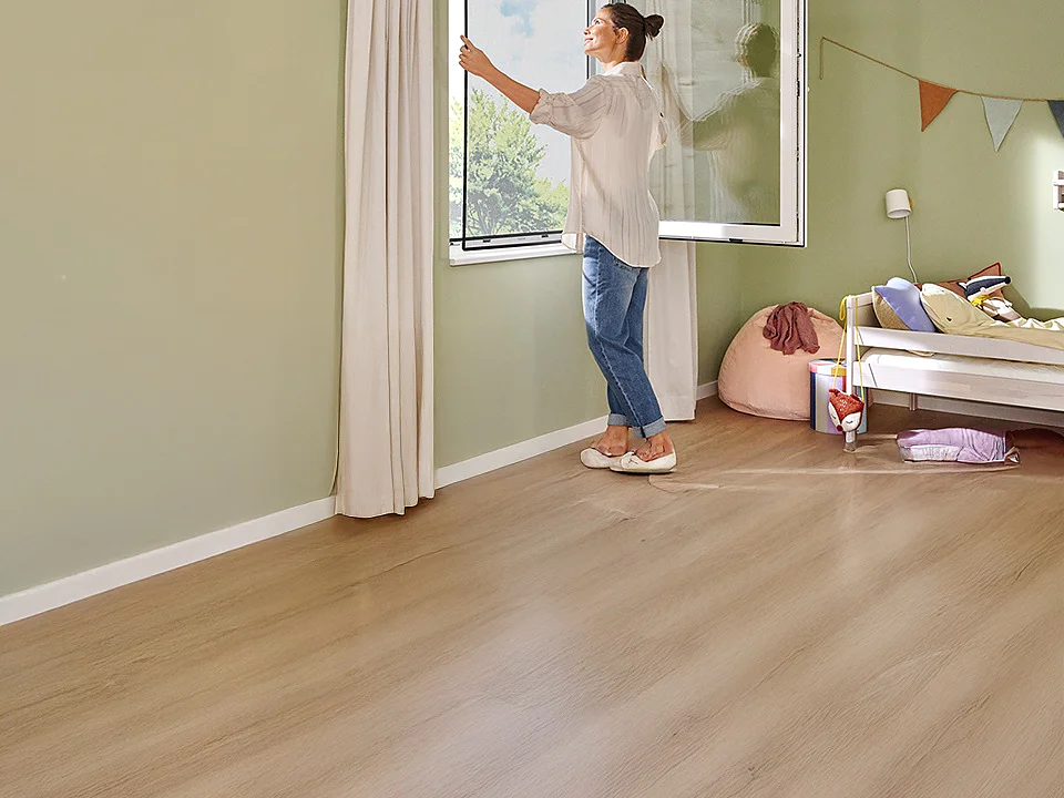 Una mujer está de pie junto a una ventana abierta en una habitación verde claro con suelo de madera. La habitación cuenta con una cama con ropa de cama y almohadas de colores vivos, así como decoraciones en las paredes fijadas con cinta adhesiva. La luz del sol entra a raudales por la ventana. (This text has been generated by AI)