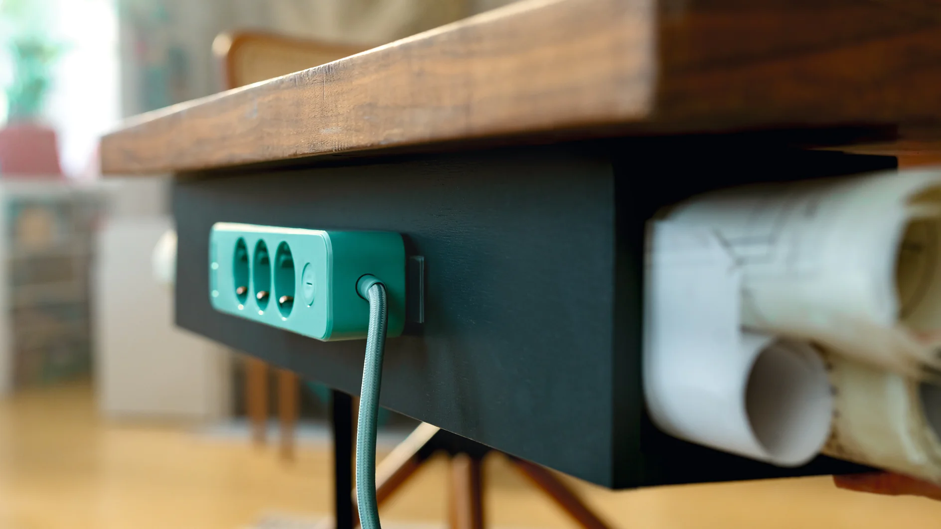 Una regleta verde azulado está montada en la parte inferior de un escritorio de madera oscura con cinta adhesiva Tesa, con un cable estampado enchufado. Los papeles enrollados se guardan en un compartimento lateral del escritorio. El fondo está suavemente difuminado. (This text has been generated by AI)